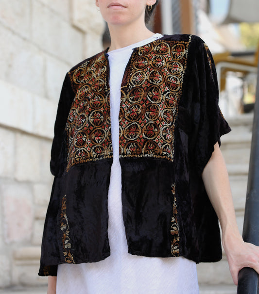 A person wearing a black velvet short-sleeve jacket with ornate multicolor embroidered panels on the front, styled over a white top in an outdoor setting.