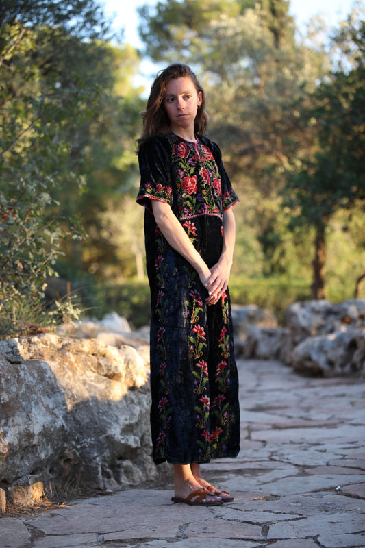 A woman stands on a stone path in a garden wearing a long black velvet dress with red and green floral embroidery and short sleeves.