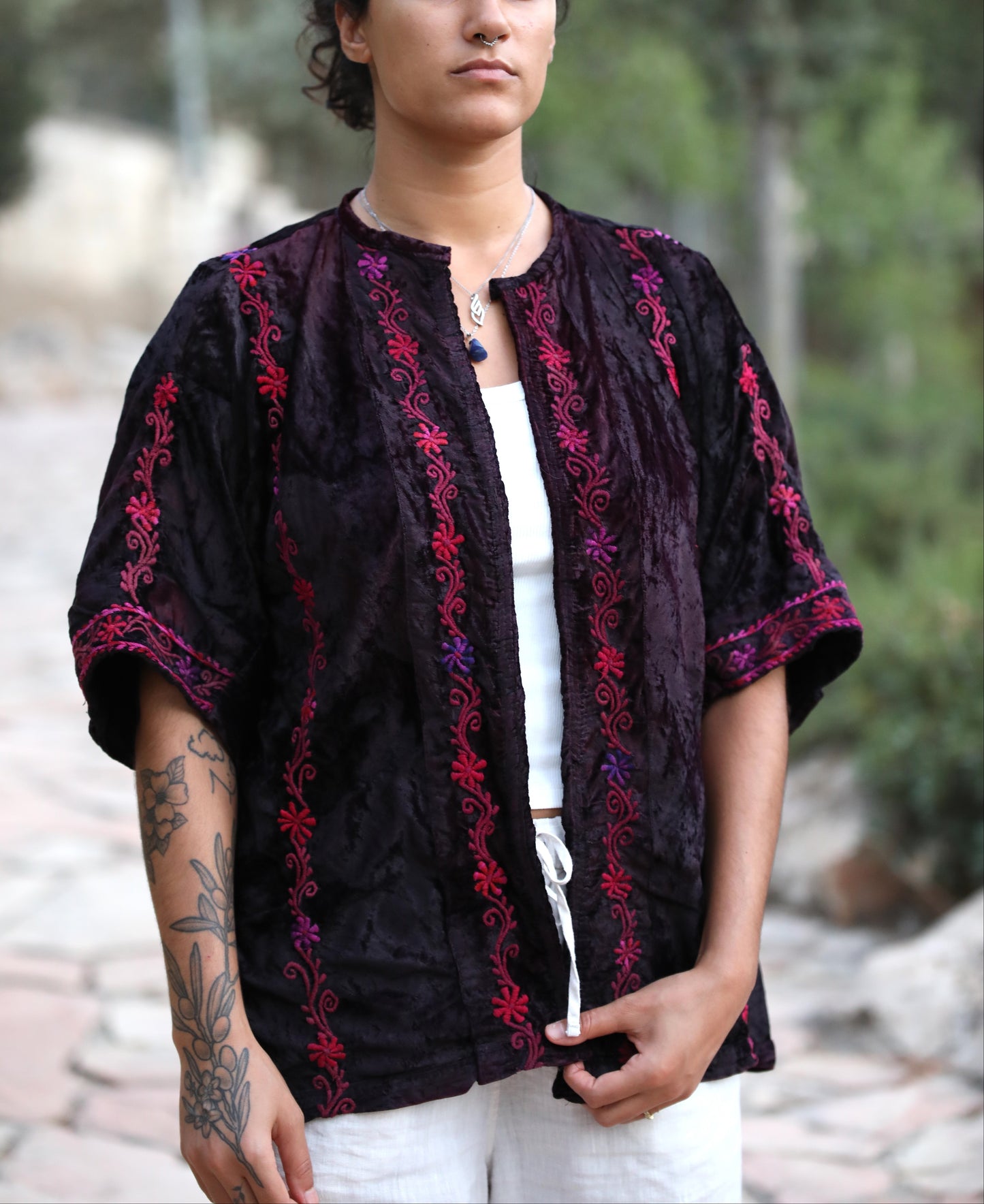 A woman wearing a black embroidered short-sleeve tunic top with red and purple floral patterns, styled over a white tank top and white pants, standing outdoors on a stone path with trees in the background.