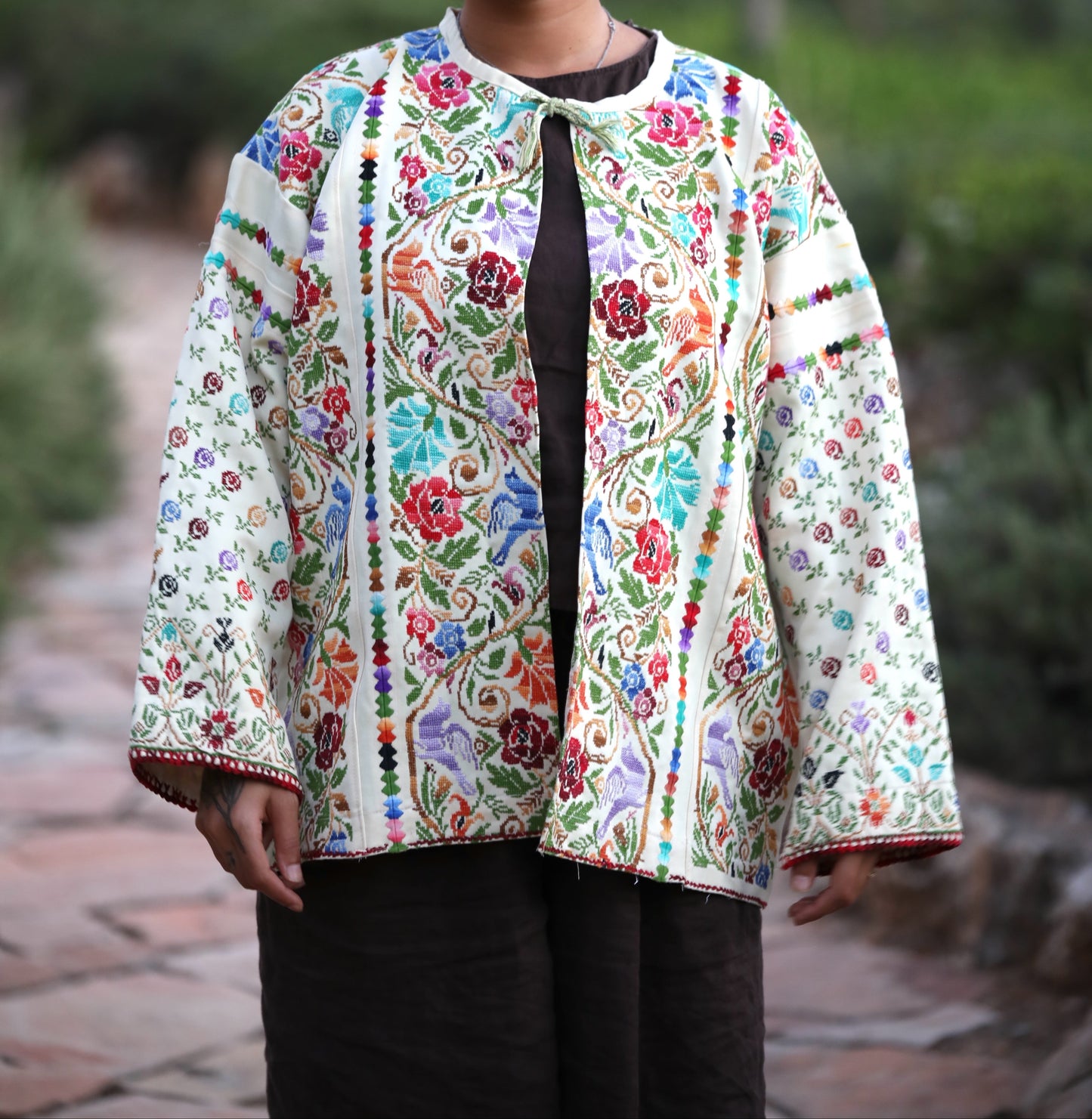 A woman wearing a colorful, heavily embroidered traditional-style jacket with a white base and vibrant floral patterns in red, blue, green, and yellow, standing outdoors on a stone path with a blurred green garden background.